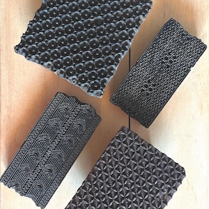 May include: Four dark brown rectangular wooden printing blocks featuring distinct carved patterns. One block displays a repeating circular motif, another a maze-like grid, a third shows crescent shapes, and the fourth features a diamond lattice design.