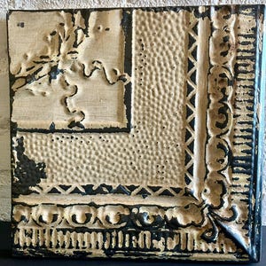 May include: Square, textured metal wall art with a distressed, vintage appearance. The piece features a cream-colored background with black detailing, including ornate borders and a floral design in the upper left corner. The overall aesthetic is rustic and decorative.