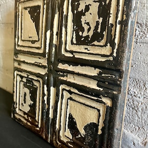 May include: Square, antique ceiling tile with a distressed, aged appearance. The tile features a repeating pattern of squares within squares, painted in black and off-white. The paint is chipped, revealing the underlying metal.