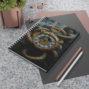 May include: Spiral-bound notebook with a detailed illustration of a gold pocket watch entwined with a golden snake against a dark, smoky backdrop. The watch face has Roman numerals. Two pencils are on the surface.