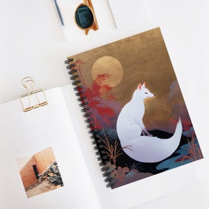 Könnte beinhalten: Ein Spiralnotizbuch mit einer Illustration eines weißen Fuchses vor einem goldenen Hintergrund mit Vollmond und rotem Laub. Das Notizbuch hat eine schwarze Spiralbindung und ist offen, mit einer leeren Seite.