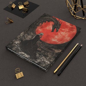 Pode incluir: Um diário de capa dura com uma ilustração de fantasia de um dragão, uma figura e uma lua vermelha. A arte da capa é predominantemente preta, cinza e vermelha. Clipes e lápis dourados são dispostos ao redor do diário.
