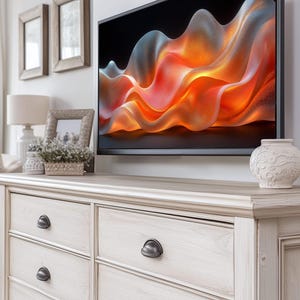 Könnte beinhalten: Eine cremefarbene Kommode mit sechs Schubladen und dunklen Metallgriffen. Ein Flachbildfernseher zeigt ein abstraktes orange-blaues Design. Dekorative Elemente sind eine kleine Vase, eine Lampe und Bilderrahmen.