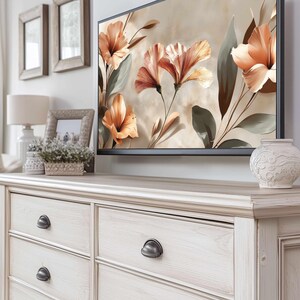 Könnte beinhalten: Eine hellfarbene Holzkommode mit sechs Schubladen und dunklen Metallgriffen. Ein großer Flachbildfernseher zeigt ein Blumenbild. Dekorative Elemente sind eine kleine Vase und gerahmte Fotos. Der Raum hat eine neutrale Farbpalette.