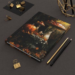 Könnte beinhalten: Ein Hardcover-Notizbuch mit einer herbstlichen Illustration mit einem Kürbis, Pilzen und Herbstblättern. Die Cover-Art ist detailliert mit warmen Farben. Goldene und schwarze Bleistifte und Büroklammern liegen auf der dunkelgrauen Oberfläche.