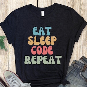 Könnte beinhalten: Ein schwarzes T-Shirt mit dem Text "Eat Sleep Code Repeat" in bunten Retro-Lettern.