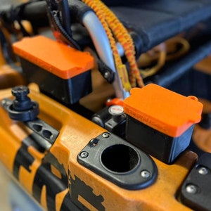 May include: Two orange and black storage containers mounted on a yellow and black kayak. The containers have lids and are secured to the kayak with screws.