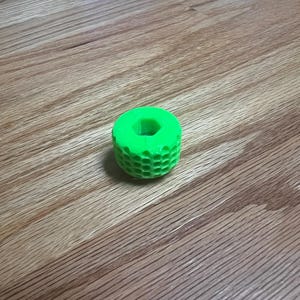 May include: A bright green, cylindrical object with a honeycomb pattern around its sides. The object has a hole through the center and is sitting on a wooden surface.