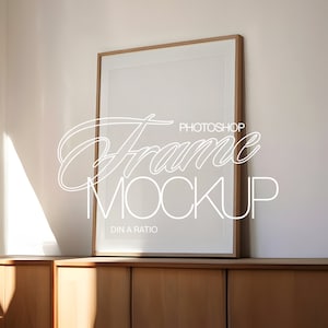 Puede incluir: Una maqueta de marco de imagen de madera con un passe-partout blanco y un fondo blanco. El marco está sentado en un gabinete de madera con un acabado de madera clara. El texto "Frame Mockup" está escrito en blanco en el marco.