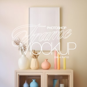 May include: A white picture frame mockup with a white mat, hanging on a light beige wall. The frame is surrounded by decorative items, including three vases with dried flowers and three candles in various pastel colors. The vases are on a light wood cabinet with glass doors.