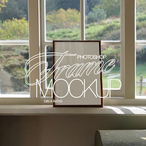 May include: A brown wooden frame with a white mat and a blank white space inside. The frame is sitting on a white windowsill with a view of green trees and grass outside the window. The text "PHOTOSHOP FRAME MOCKUP DIN A RATIO" is written in white on the frame.