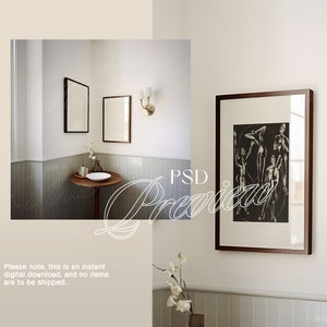 Op de afbeelding: Een badkamerinterieur met witte muren en grijze betegelde vloeren. Twee ingelijste spiegels hangen aan de muur, een boven een wastafel en de andere in de hoek. Een ingelijste zwart-witprint van een figuurtekening hangt aan de muur boven een klein tafeltje met een witte keramische wastafel. Een kleine plant staat in een bruine pot op de tafel. Een gouden wandlamp met een witte kap bevindt zich aan de muur rechts van de spiegels.