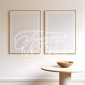 May include: Two empty picture frames with a light wood frame, hanging on a white wall. A round white table with a wooden bowl filled with fruit sits in front of the frames.