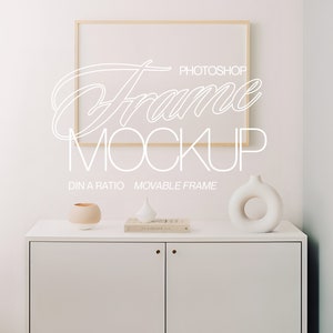 May include: A white cabinet with two doors and gold knobs. A wooden frame with a white mat is hanging on the wall above the cabinet. There are three decorative objects on top of the cabinet: a white vase, a small white ceramic object, and a brown ceramic pot. The text "Frame Mockup" is written in white on the wall.