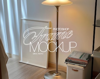 May include: A white picture frame mockup leaning against a wall, with the text "Frame Mockup" in a cursive font. A tall, bronze floor lamp with a round shade stands nearby. A stack of magazines rests on a white storage box.