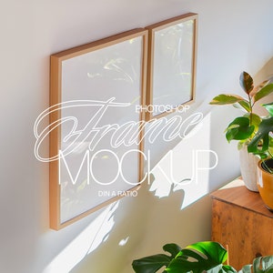 May include: Two wooden picture frames with white mats hanging on a white wall. The frames are in a living room setting with plants in pots on a wooden table.