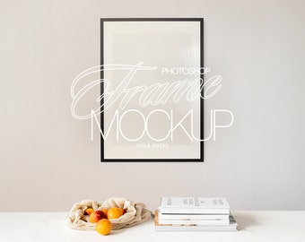 Op de afbeelding: Een zwarte fotolijst mockup met een witte achtergrond, hangend aan een lichtgrijze muur. Een witte mesh tas gevuld met perziken staat op een witte tafel met een stapel boeken.