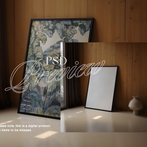 May include: A black framed picture with a watercolor painting of a forest scene. A smaller black framed picture is on the floor in front of the larger picture. A white vase is on the floor to the right of the smaller picture.