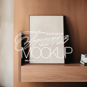 May include: A white framed picture mockup on a wooden shelf with a stack of books on the right side. The books have titles that include "Just Me" and "The Old Man and the Sea".