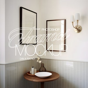 Op de afbeelding: Twee ingelijste foto's hangen aan een witte muur in een badkamer. De lijsten zijn van zwart en bruin hout. Een kleine tafel met een witte kom en flessen staat in de hoek van de kamer.