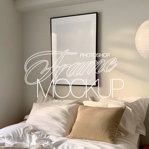 May include: A black picture frame mockup on a white wall above a bed with white bedding and a tan pillow. A white paper lantern hangs from the ceiling.