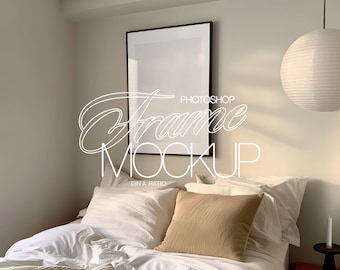 May include: A black picture frame mockup on a white wall above a bed with white bedding and a tan pillow. A white paper lantern hangs from the ceiling.