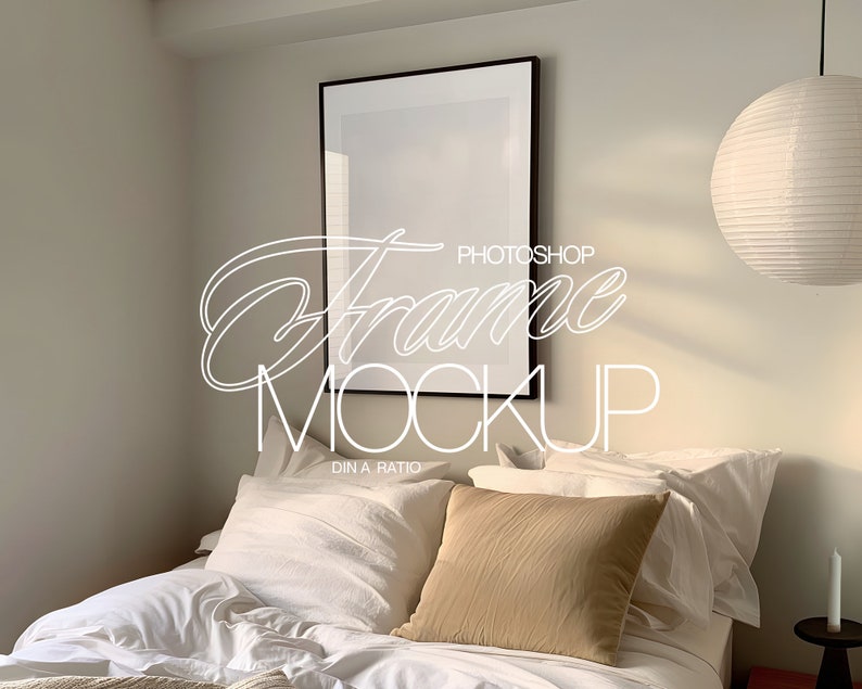 May include: A black picture frame mockup on a white wall above a bed with white bedding and a tan pillow. A white paper lantern hangs from the ceiling.