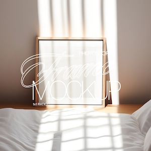 May include: A wooden frame with a white mat and a white background. The frame is sitting on a wooden table next to a white bed with white bedding.
