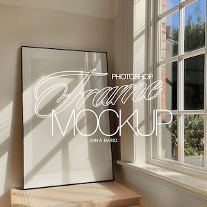 May include: A black picture frame mockup with a white mat and a white background. The frame is sitting on a light brown table with a woven texture. The frame is in front of a window with white trim and a view of green trees and a blue sky.