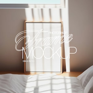 May include: A wooden frame with a white mount and a blank white canvas inside. The frame is sitting on a wooden table in front of a white wall with a window behind it. The frame is in a bedroom setting with a white bed and pillows in the background.