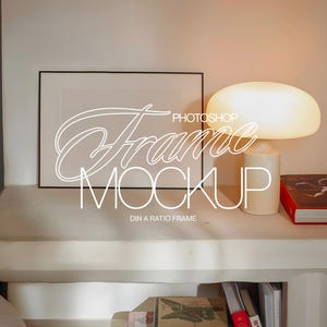 以下が含まれることがあります： 「Photoshop Frame Mockup」と「Din A Ratio Frame」の文字が入った黒いフレームモックアップ。白いシェードの白いランプと赤い本が白い棚に置かれています。棚の下には本が見えます。