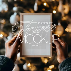 May include: A person holds a brown wooden picture frame with a white mat and a blank white space inside. The frame is a mockup for a photo or artwork. The text "Photoshop Frame Mockup DIN A Ratio" is printed on the frame.