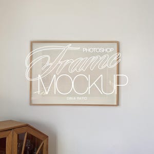 Könnte beinhalten: Ein rechteckiger Holzrahmen-Mockup mit dem Text "Photoshop Frame Mockup" in weißer, kursiver Schrift. Der Rahmen hängt an einer weißen Wand über einem Holzschrank. Das Bild hat eine neutrale, minimalistische Ästhetik.
