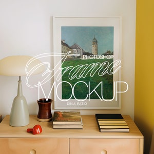 May include: A white picture frame with a landscape painting of a town with a tall tower. The frame is hanging on a white wall above a wooden dresser with a lamp, a ceramic vase, and books. The dresser is next to a yellow wall with a brown curtain.
