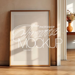 May include: A gold-coloured frame with a white mount and a white background. The frame is sitting on a wooden floor with a light brown wall behind it. The text "Photoshop Frame Mockup DIN A Ratio" is visible in the frame.