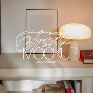 May include: A white lamp with a round shade sits on a white shelf with a black frame and a book. The frame is empty and the text "Photoshop Frame Mockup" is written in white on the image. The text "DIN A RATIO FRAME" is written in white below the text "Photoshop Frame Mockup".