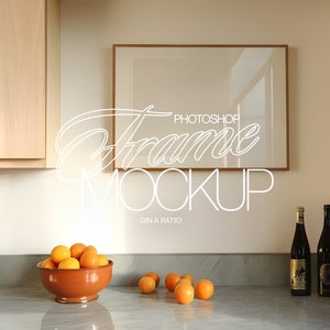 Puede incluir: Una maqueta de marco de imagen con un marco de madera colgado en una pared beige claro sobre una encimera de cocina. Hay dos botellas de vino y un tazón de naranjas en la encimera.