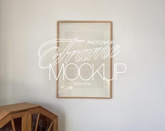 May include: A rectangular wooden frame mockup with a white background. The frame contains a white interior with the text "Photoshop Frame Mockup" in a cursive font. A wooden cabinet is visible in the bottom left corner.