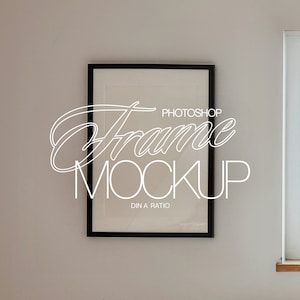 May include: A black picture frame mockup hanging on a white wall with a window to the right. The frame is empty and has a white border. The text "Photoshop Frame Mockup DIN A Ratio" is written in white on the frame.