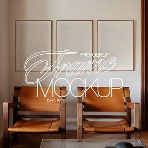 May include: Three empty picture frames with a wooden frame and a white mat are hanging on a white wall. The frames are arranged in a row, with the middle frame slightly higher than the other two. The frames are in a living room setting with two brown leather chairs and a wooden floor.