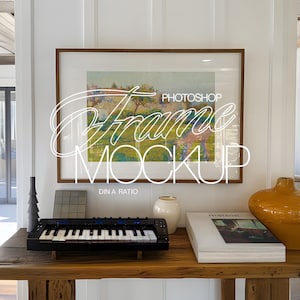 Puede incluir: Una consola de madera exhibe una obra de arte enmarcada, un teclado pequeño, objetos decorativos y una pila de libros. La obra de arte es una pintura de paisaje. El texto "Photoshop Frame Mockup" está superpuesto en la imagen.