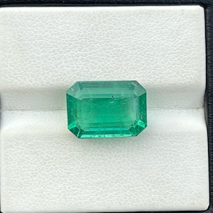 May include: A vibrant green emerald gemstone, cut in an emerald shape, is displayed in a white ring box.