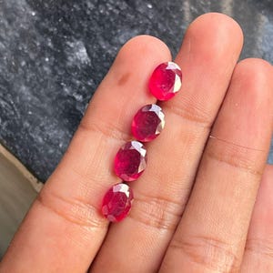 Könnte beinhalten: Vier oval geschliffene, facettierte Rubin-Edelsteine. Die Rubine haben eine tiefe, leuchtende rote Farbe und werden in einer Hand präsentiert. Die Edelsteine sind in einer vertikalen Linie angeordnet und zeigen ihre polierten Oberflächen und ihre innere Klarheit.