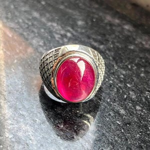 Può includere: Un anello color argento con una grande pietra preziosa ovale di un vibrante rosso rosato incastonata al centro. La fascia in metallo presenta una dettagliata trama a griglia in rilievo che si estende lungo i lati della montatura.
