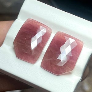 May include: Two faceted, rectangular gemstones with a pinkish-red hue. The gems have a geometric cut with multiple facets that reflect light. They are displayed on a white surface.
