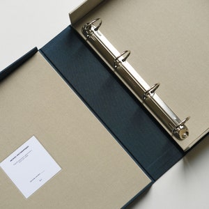 May include: A navy blue and beige binder, open to reveal a three-ring metal mechanism. The interior has a beige surface with a navy blue spine. A small white label with text is visible. Designed for archival storage, the binder is well-suited for documents.