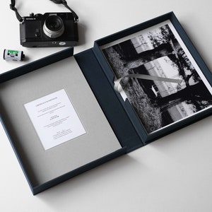 May include: A navy blue presentation box with a gray interior holds a black and white photograph. A black camera and film canister are placed nearby. A certificate is visible inside the box. The photograph features a landscape scene.