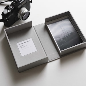 Könnte beinhalten: Eine graue Präsentationsbox mit einem Schwarz-Weiß-Foto. Die Box ist geöffnet und zeigt das Foto und ein Echtheitszertifikat. Im Hintergrund ist eine schwarze Kamera mit Objektiv zu sehen.