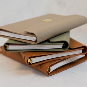Può includere: Una pila di tre diari rilegati in pelle di colore beige, verde e marrone. I diari hanno anelli dorati e sono chiusi.