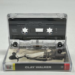 May include: A clear cassette tape featuring Clay Walker's album. The tape has a black label with white text listing song titles like "Dreaming With My Eyes Open." The cassette case is open, revealing the tape and album art. The album art features a black and white image of a man.